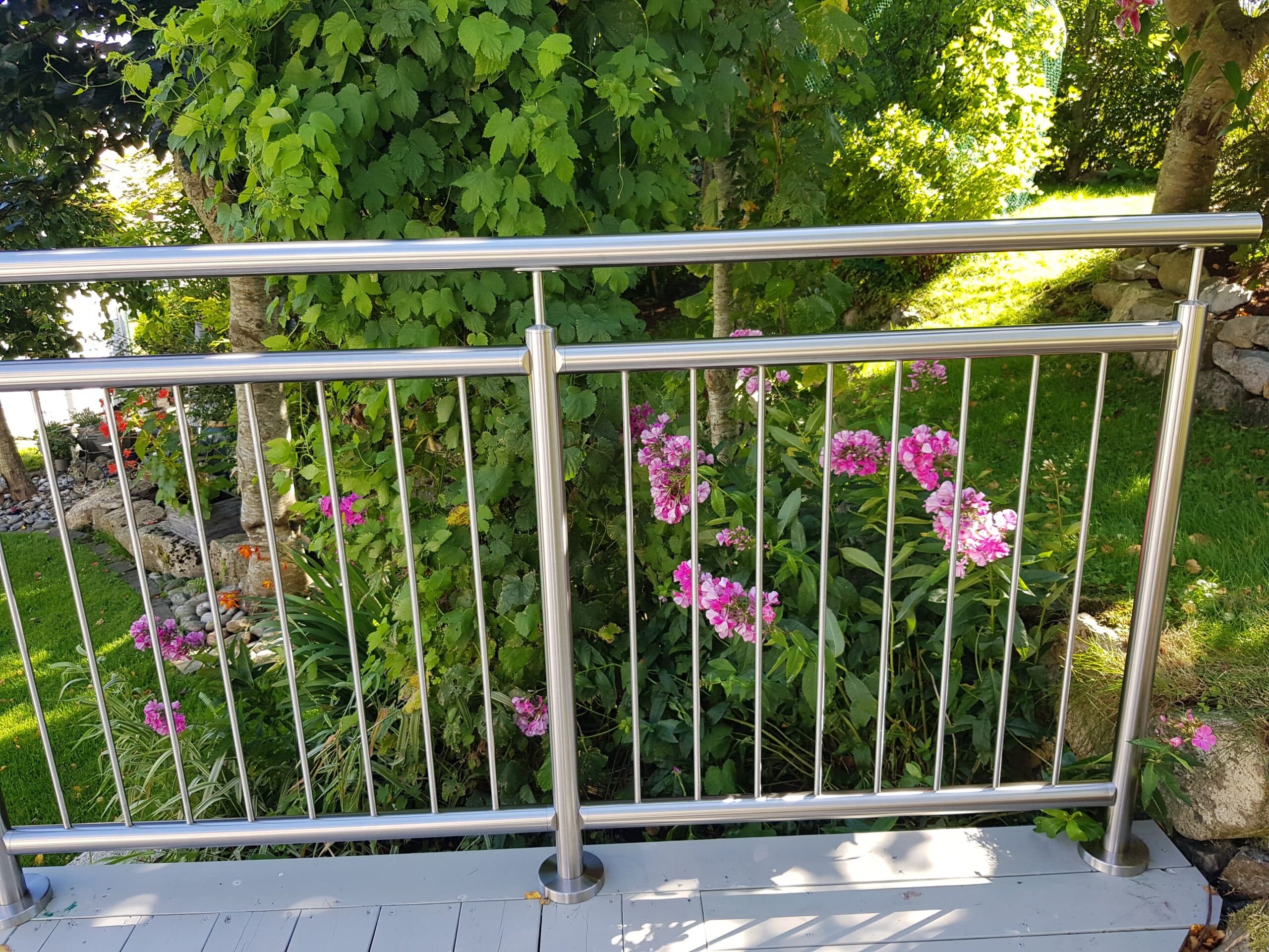Garde-corps vertical en inox installé sur terrasse avec fleurs colorées et vue sur un jardin verdoyant en arrière-plan.