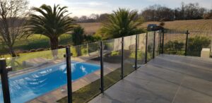 Glass Pool Railing with Black Frame and Unobstructed View Modern glass pool fence with laminated clear glass and black aluminium posts surrounding a palm-lined pool area with open landscape views.