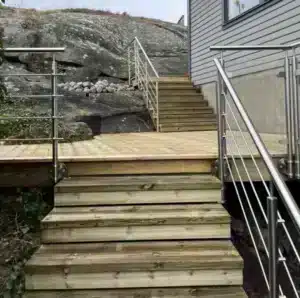 Rostfritt räcke med horisontella ståndare Trappa och altan i tryckimpregnerat trä med rostfritt räcke. Räcket har horisontella ståndare och monteras längs trappor och avsatser. Huset har en fasad i grå panel och berg i bakgrunden.