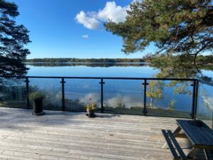 Sort glasværn med søudsigt Træterrasse med sort stolpeglasværn, der indrammer en grund ved søen og giver frit udsyn over vand og natur.