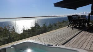 Pfostenfreies Glasgeländer auf einer Terrasse am Meer mit Holzboden und Gartenmöbeln im Schatten einer Markise.