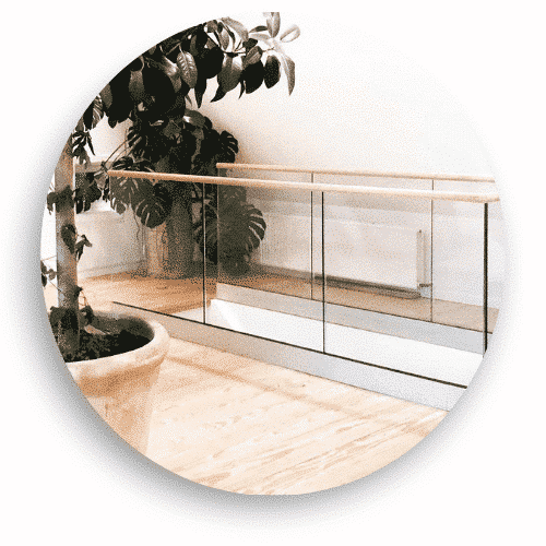 Ett glasräcke med klämfästen och en överliggare i trä, installerat inomhus längs ett loft. Räcket kombinerar transparent glas och varma trädetaljer för en luftig och säker avgränsning i rummet. Grönska och ljus träparkett kompletterar den naturliga känslan.