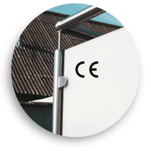 ärbild av ett CE-märkt vindskydd med opalt glas och rostfria stolpar. Räcket har en rund överliggare i rostfritt stål och glaset är fastsatt med klämfästen. I bakgrunden syns ett tegeltak och en blå himmel, vilket framhäver räckets stilrena och funktionella design.