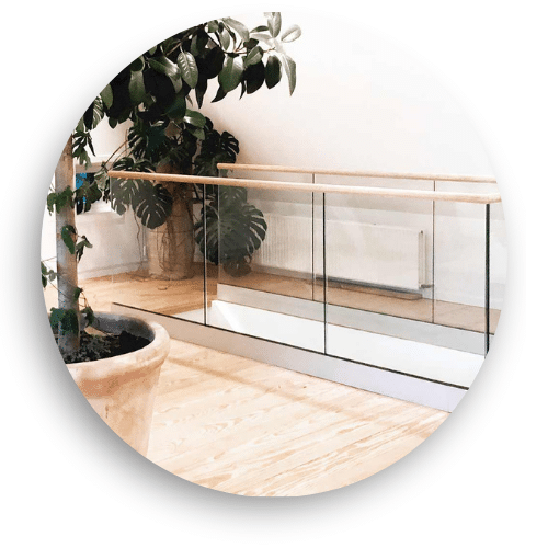 Garde-corps intérieur en verre avec main courante en bois dans une pièce claire avec plancher bois et plantes vertes.