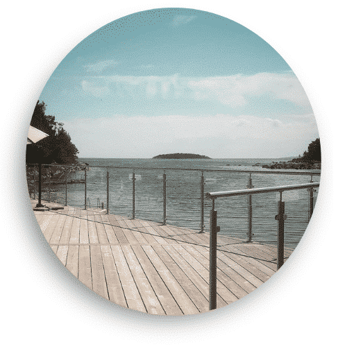 Glasräcke med rostfria stolpar monterat längs en träbrygga vid vattnet. Räcket ger en säker och diskret inramning utan att störa den öppna utsikten över havet och horisonten.