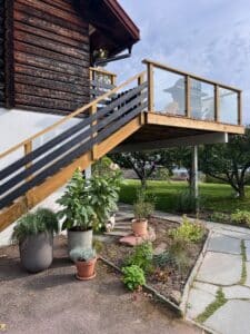 Hochgelegene Terrasse mit Holz- und Klarglasgeländer an einer Blockhausfassade, mit Blick auf Garten und Grünflächen.