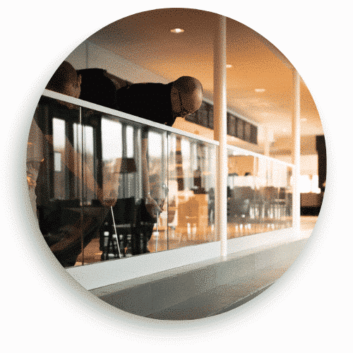 Två montörer arbetar med installationen av ett glasräcke med vit överliggare i en ljus offentlig miljö. Räcket har klämfästen och glaset reflekterar omgivningen. I bakgrunden syns en restaurang eller café med bord och stolar.