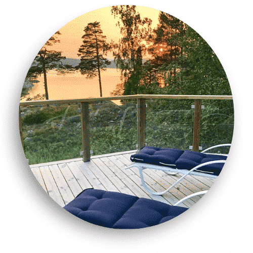 Trämonterat glasräcke med klämfästen omger en altan med utsikt över en sjö vid solnedgång. Stolpar i trä och en överliggare ramar in de klara glasskivorna, vilket skapar en säker men öppen känsla mot naturen. Två solstolar med mörka dynor står placerade på trädäcket.