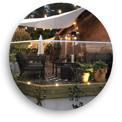 Holzterrasse mit pfostenfreiem Glasgeländer, Gartenmöbeln, Pflanzen und stimmungsvoller Beleuchtung.