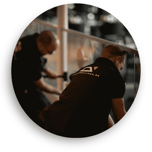 Två montörer i svarta arbetskläder installerar ett glasräcke i en offentlig miljö. De arbetar noggrant med justering och montering av glaset, som hålls på plats av rostfria fästen. Reflektioner i glaset ger en känsla av rymd och transparens i lokalen.