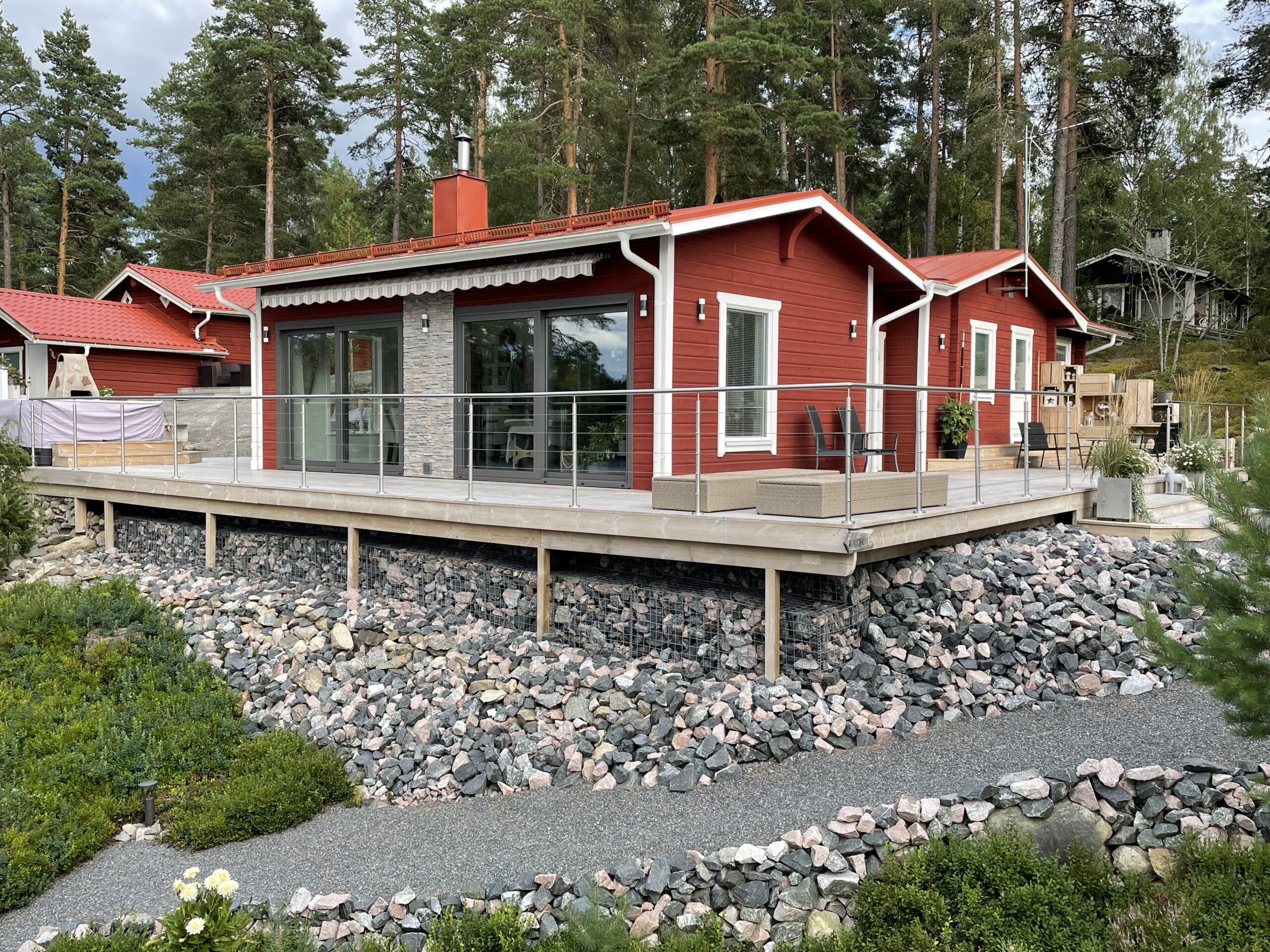 Edelstahl-Seilgeländer rund um große Terrasse eines roten Holzhauses in Küstenlandschaft – perfektes Geländer für Höhenunterschiede und freie Sicht in den Garten.