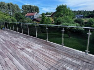 Balkon¬geländer mit getöntem Sicherheitsglas und Edelstahlpfosten auf Holzterrasse mit Blick auf Garten und Haus.