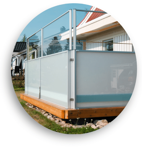 Höj- och sänkbart glasräcke med aluminiumstolpar och en kombination av klart och frostat glas, monterat på en upphöjd träterrass vid ett vitt småhus.