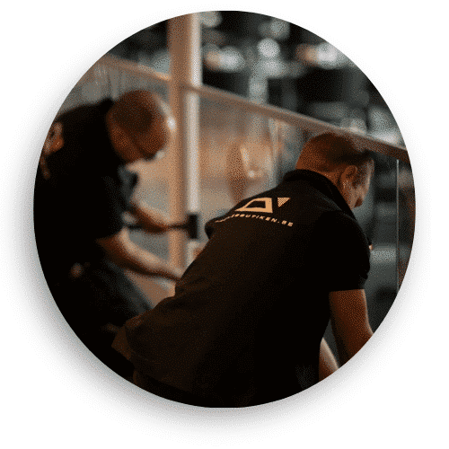 Zwei Monteure von Geländerladen arbeiten an der Montage eines Glasgeländers. Sie tragen schwarze Arbeitskleidung mit Firmenlogo und passen Details für einen sicheren Sitz an. Das Glas spiegelt die Umgebung im Innenbereich wider