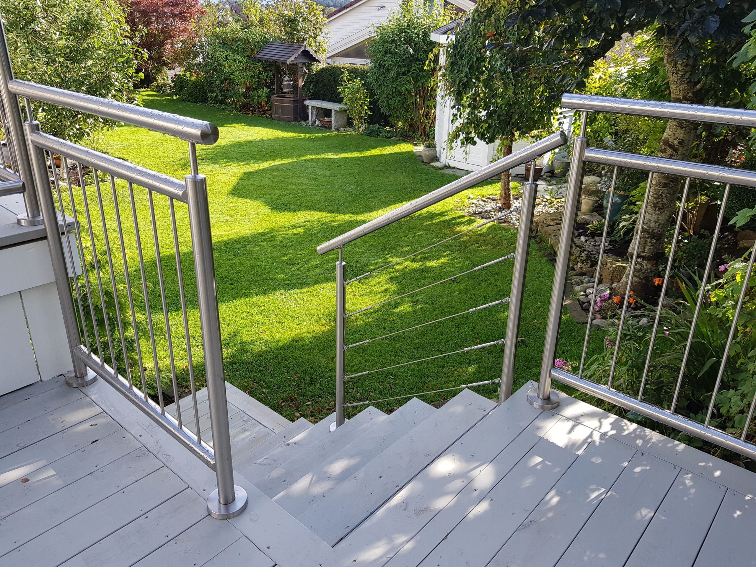 Escalier depuis une terrasse équipé d’un garde-corps en inox avec câbles, entouré de pelouse, arbustes et massifs fleuris.
