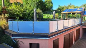 Terassi huurrelasikaiteella ja pyöreillä ruostumattomilla tolpilla, ympärillä vehreä puutarha ja ulkohuone