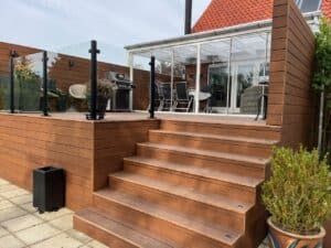 Hævet altan med sort værn Højtliggende altan med sort aluminiumsrækværk og klart glas, omgivet af brun terrasse og trappe i flere niveauer.