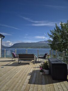 Große Terrasse mit Glasgeländer und Holzpfosten, gelegen am See mit Panoramablick und stilvoller Außenmöblierung.