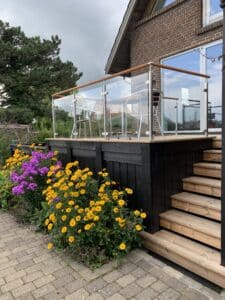 Blomstrende terrasse med træhåndliste Altanværn i rustfrit stål og klart glas med træhåndliste, sort træbeklædning og farverige blomster i forgrunden.