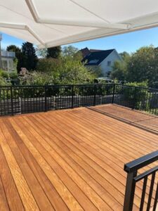 Sort altanrækværk i smedejern omkring terrasse i træ med markise, grøn beplantning og solrigt udemiljø.