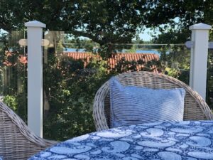 Nahaufnahme einer Terrasse mit weißem Aluminiumgeländer und Klarglas, Rattanmöbeln, blauen Textilien und Seeblick.