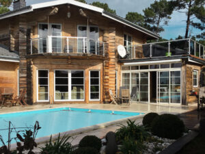 Glass railing overlooking pool and garden Timber villa with glass railing surrounding upper balcony and lower terrace, with pool and pine trees in garden setting.
