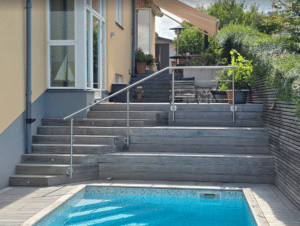 Stainless steel stair railing beside pool area Outdoor stainless steel handrail mounted by wide wooden pool steps, complementing adjacent glass fencing for full perimeter safety.