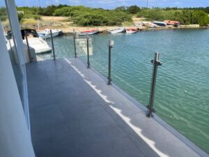 Glass railing by mooring with stainless steel posts Glass and stainless steel balustrade along dock overlooking small marina, with trees and calm water in background.