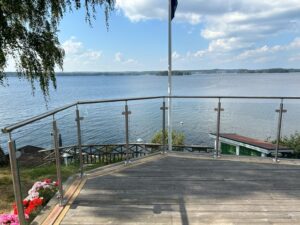 Stainless steel glass railing with lake view Timber deck with round stainless steel post and clear glass balustrade, facing lake and wooden jetty below.