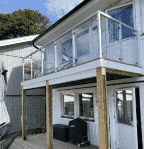 Glasräcke med fyrkantiga aluminiumstolpar i vitt monterat på balkong med trall och husfasad i trä