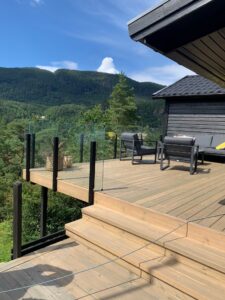 Frameless black aluminium balustrade with forest view Deck with sleek black aluminium frameless glass railing, offering views of trees, hills and open blue sky.