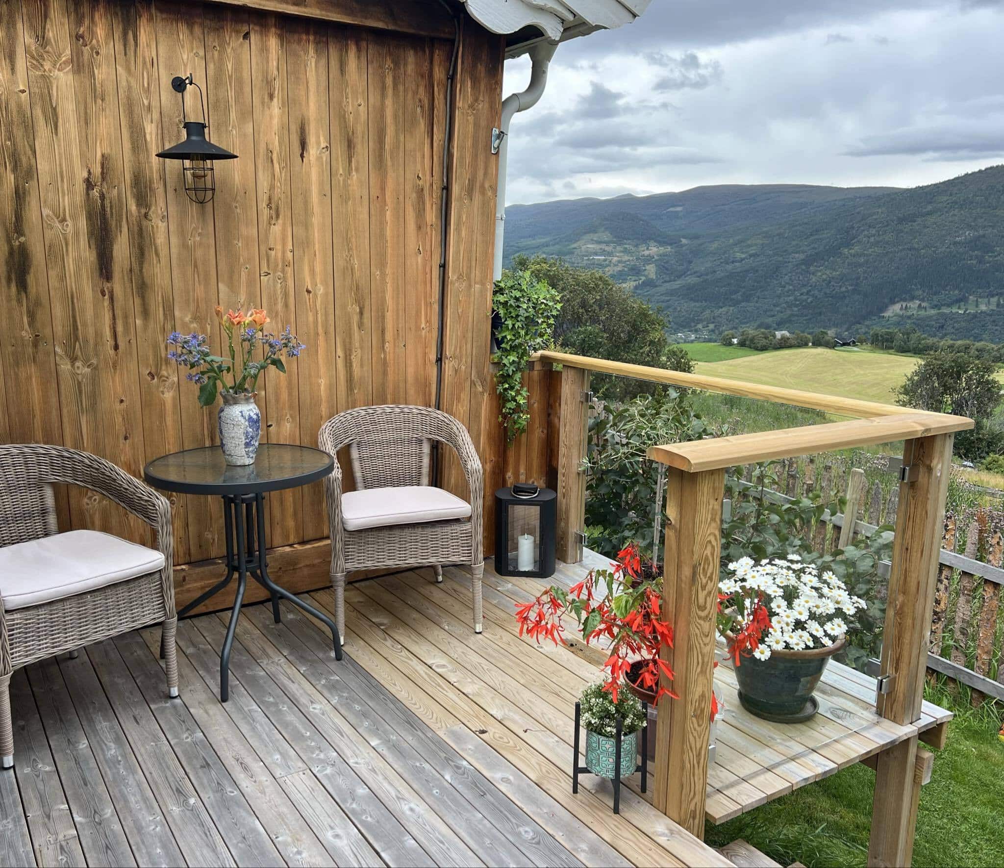 Holzterrasse mit Glasgeländer, Blick auf Tal und Felder im Hintergrund. Zwei Stühle und ein Café-Tisch stehen an der Hauswand.
