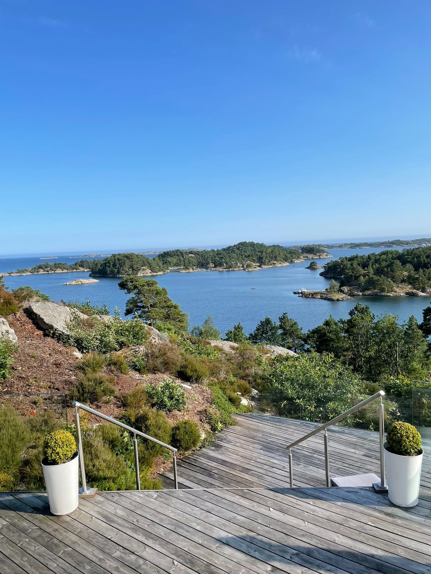 Stainless steel wire handrails on wooden deck stairs framed by potted plants, leading down towards a coastal hillside with islands and open sea.