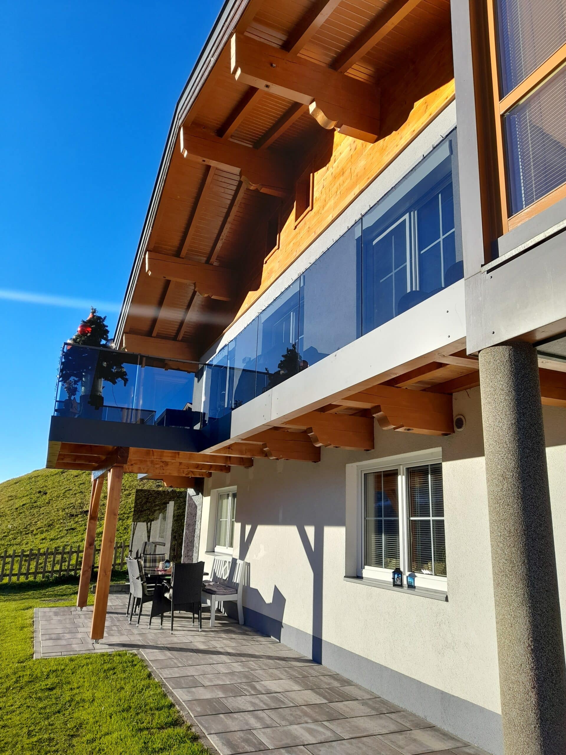 Kehyksetön lasikaide modernin puualppimajan parvekkeella, taustalla vuoristonäkymä ja alapuolella patiopiha.