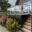 Glasbrüstung mit Holzhandlauf und Blumenbeet Geländer mit Edelstahlpfosten, Glas und rundem Holzhandlauf bei schwarzer Terrasse mit bunten Blumen entlang der Mauer.