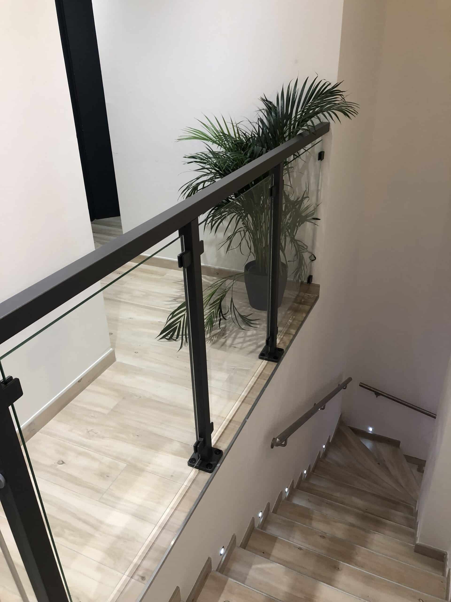 Schwarzes Aluminiumgeländer mit Klarglas, montiert entlang einer Treppe im Innenbereich mit hellem Holzfußboden und integrierter LED-Beleuchtung.