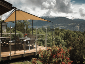 Höhenverstellbare Terrasse mit Glasgeländer, Edelstahlpfosten und Sonnensegel – auf Rädern montiert mit Blick auf grüne Täler & Alpen.