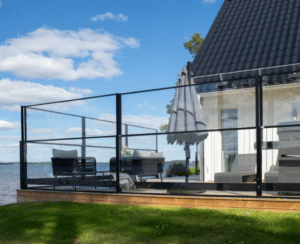 Sort aluminiumshegn med glas Udeområde med sort aluminiumshegn og store glaspaneler som læhegn. Monteret på træterrasse ved hvidt hus med sort tag. Udsigt til sø og blå himmel med skyer i baggrunden.