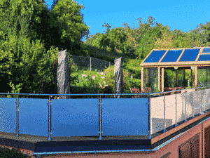 Balkongeländer aus Edelstahl mit satiniertem Glas, montiert auf einer erhöhten Terrasse. Das Geländer hat runde Pfosten und einen Handlauf aus Metall. Im Hintergrund sieht man einen grünen Garten, ein Gewächshaus mit Glasdach und einen blauen Himmel.