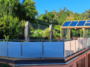 Balkongrekkverk i rustfritt stål med frostet glass, montert på hevet terrasse. Rekkverket har runde stolper og overligger i metall. I bakgrunnen ses grønn hage, drivhus med glasstak og blå himmel.