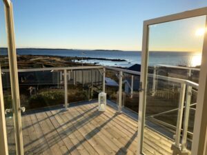 Balkon mit Glasgeländer und weißem Aluminiumrahmen, offenen Flügeltüren und Sonnenuntergang über dem Meer und den Schären im Hintergrund.