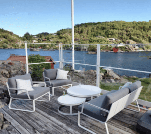 White aluminium glass balustrade with sea view Cliffside terrace with clear glass and white aluminium railing, grey garden furniture and views of fjord, red cabins and hills.