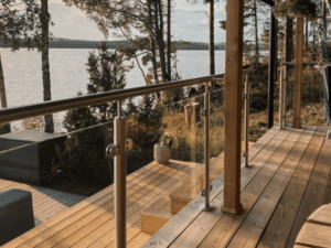 Holzterrasse mit Glasgeländer aus Edelstahlpfosten und Holzhandlauf – Blick auf See, bewaldete Ufer und Holztreppe zum Wasser.