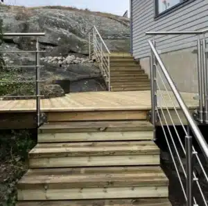 Edelstahlgeländer mit Horizontalstäben Holztreppe und Balkon mit druckimprägniertem Belag und Edelstahlgeländer mit horizontalen Stäben. Die moderne Fassade und Bergkulisse runden das Bild ab.