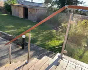 Treppen- und Balkongeländer mit Drahtseilen und Glas Außentreppe mit Geländer aus Edelstahl, Drahtseilen, Glas und Holzhandlauf. Der Garten im Hintergrund mit Rasen und Holzhaus unterstreicht das naturnahe Design.