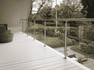 Glasräcke med rostfria stolpar och handledare på en vitmålad balkong, med utsikt över en lummig trädgård.