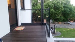Glasgeländer mit schmalem Tor auf der Terrasse Glasgeländer mit schmalem Tor auf dunklem Holzdeck am Eingang, mit schwarzem Aluminiumrahmen und Blick auf Garten und gepflasterten Sitzbereich.