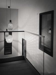 Stilvolle Minimalistik in Schwarz-Weiß Moderne Innenarchitektur mit pfostenfreiem Glasgeländer und Hängelampen, die grafische Lichtmuster auf Wände mit Holzfußboden werfen.