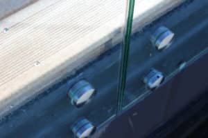 Detail einer Punkthalterung aus Edelstahl Nahaufnahme einer Punkthalterung eines pfostenfreien Glasgeländers mit Edelstahlabstandshaltern, montiert auf Holz- und Betonuntergrund.