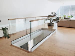 Pfostenfreies Glasgeländer an Treppe im Innenbereich Elegantes pfostenfreies Glasgeländer rund um Treppenöffnung in moderner Dachwohnung mit Holzhandlauf, weißer Sockelleiste und hellem Parkett.